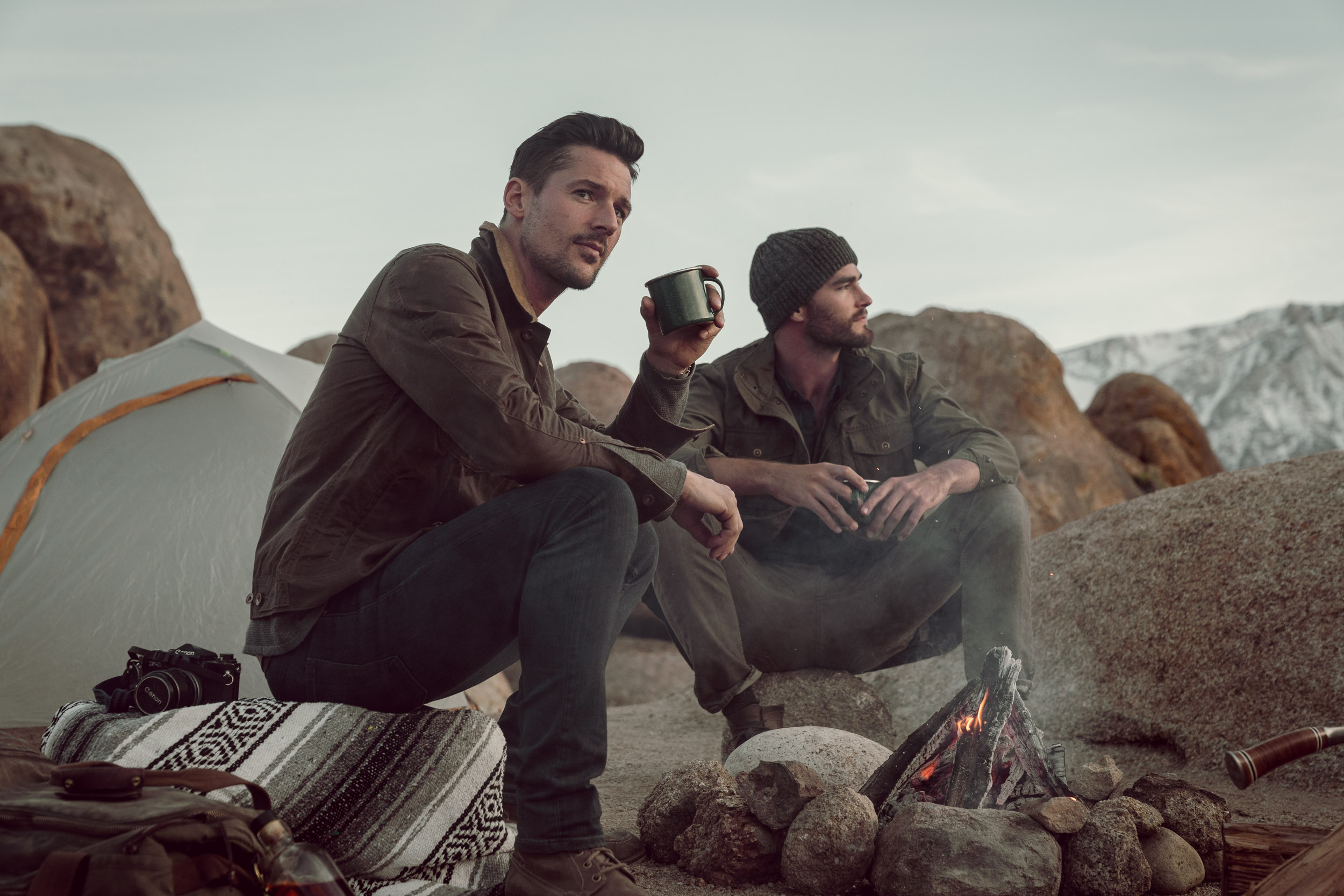 Two rugged outdoorsmen enjoying a drink by the fire at twilight.