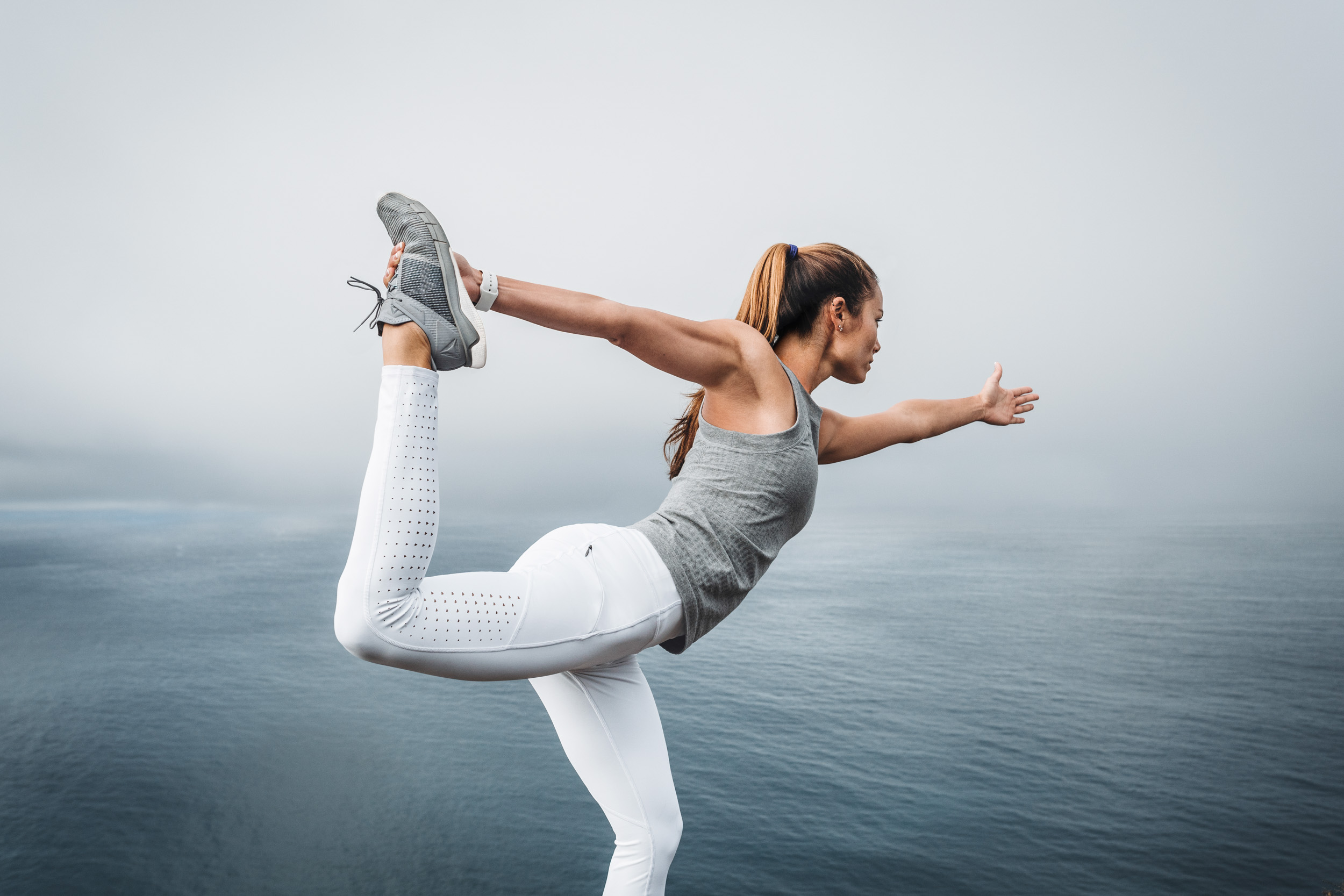 Female athlete wearing white Athleta tights and grey shirt practices yoga on a cliff near a foggy ocean