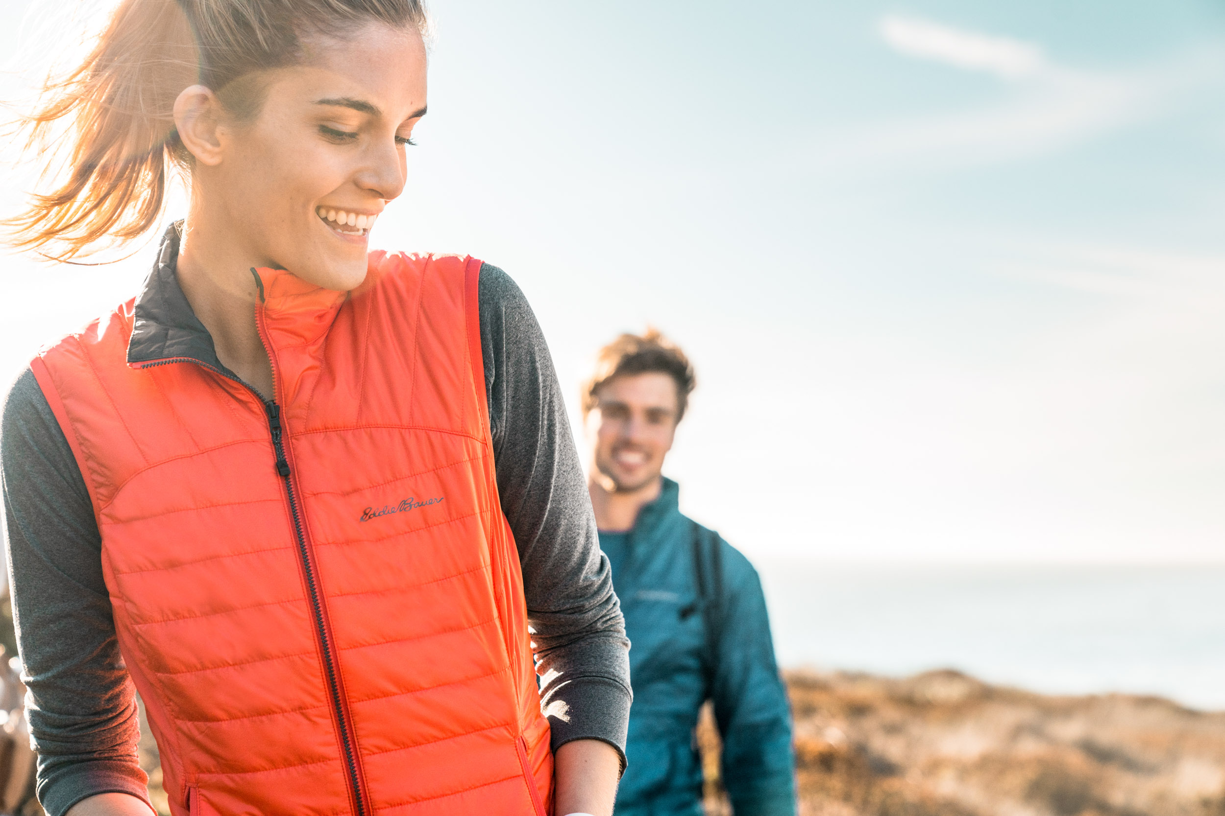 Female athlete in red vest looks back at male athlete in blue jacket on the coast