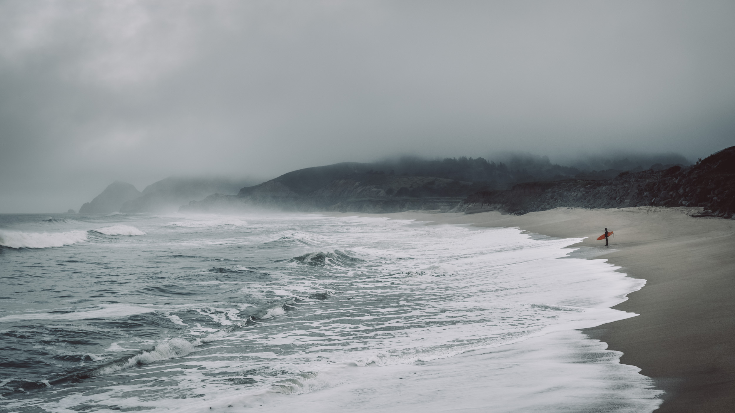 A lone male surfer with red surfboard stands on a wild and foggy beach with huge waves