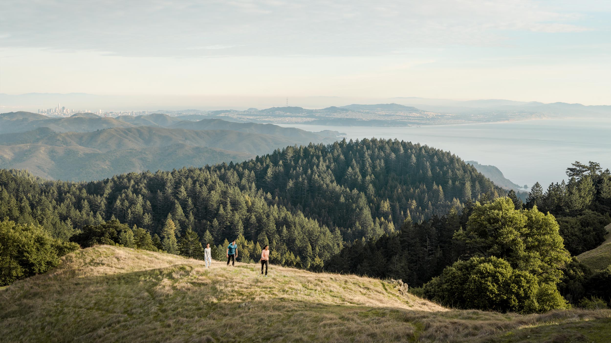 Three hikers walk across a grassy ridge line with a massive view San Francisco and the Pacific Ocean