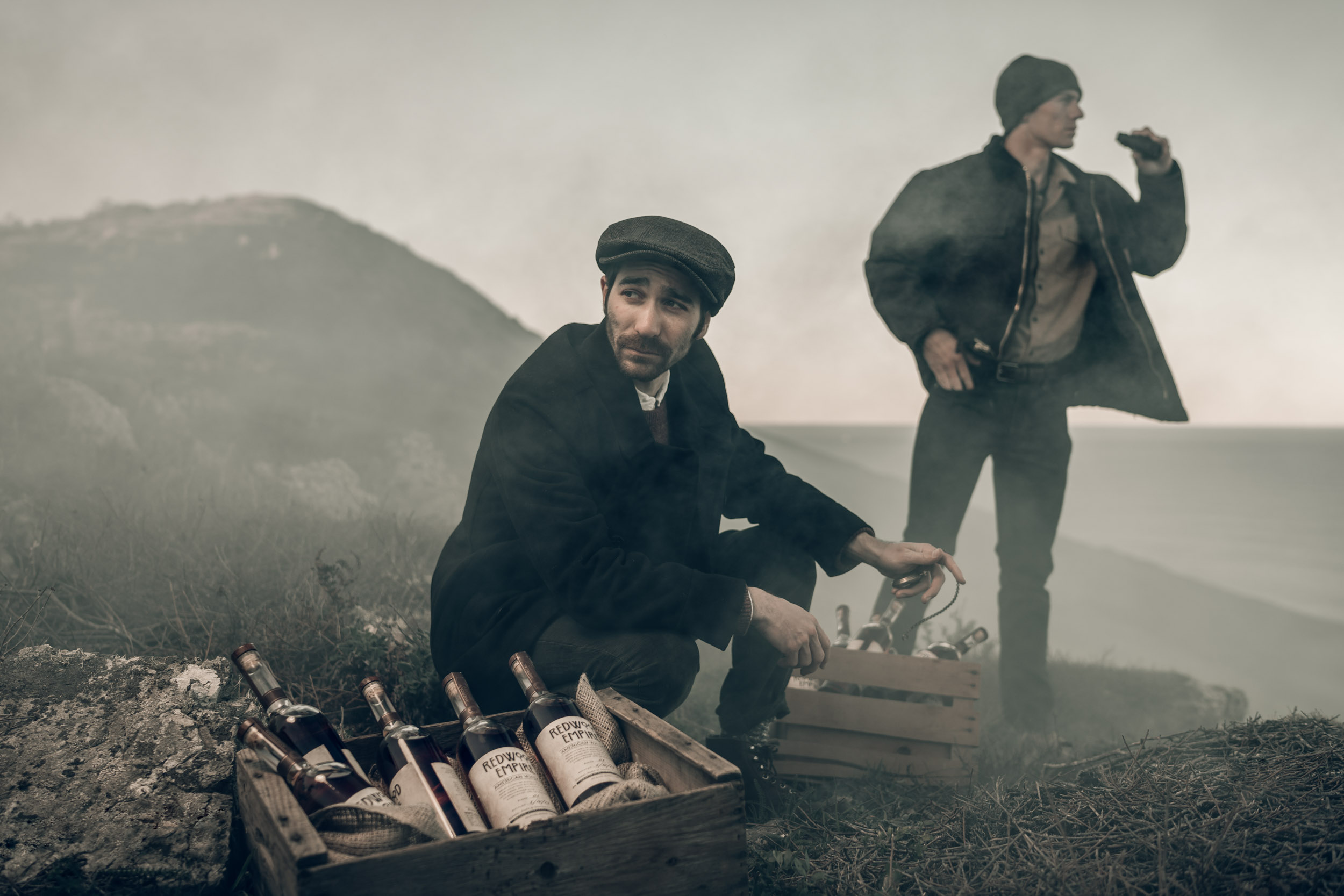 Two male bootleggers stand on the foggy coast guarding a crate of Redwood Empire whiskey bottles